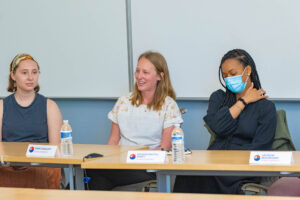 3 student panelists sit at a table in the front of a classroom and two are laughing.