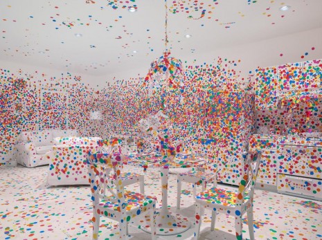 A painted white room almost entirely covered with rainbow stickers. The room has four chairs, a table, a visible couch and kitchen area. The hanging ceiling lamp is also fully covered with stickers.