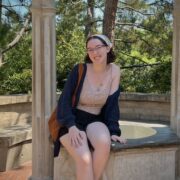 Young woman with shoulder length hair and glasses, seated on a stone bench, smiles at the camera.