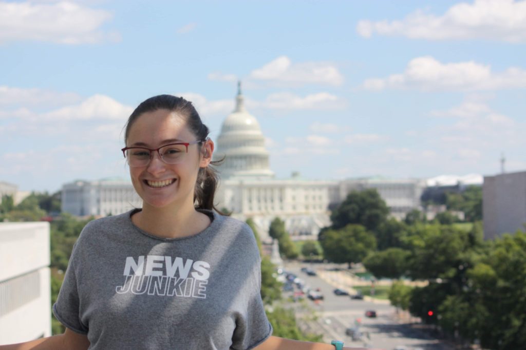 A woman in glasses stands in a tee shirt reading "News Junkie" with the Capitol building in the distant background