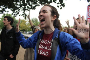 Robin at a protest, with her mouth open as she yells out a chance. Her two hands are held up in protest, with "I BELIEVE" written on them in sharpie. 