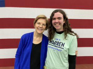 Elizabeth Warren and Robin with their arms around each other and smiling at the camera. Robin is wearing a liberty green "I'm a Warren Democrat" t-shirt, and Warren is wearing a dark blue cardigan. The background is red and white stripes of the flag.