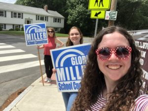 Robin is taking a selfie with two other young women in the background holding Gloria Miller for School Committee Signs. They are by a road and sidewalk. All 3 are smiling at the camera.
