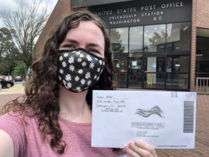Robin is standing in front of a US Postal Office. She is holding an envelope for a mail-in ballot in her hand and is taking a selfie. She is wearing a fabric mask with flowers. 
