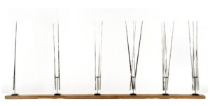 Twenty-one stainless steel blades, just over three feet tall each, rest atop six metal hinges on a long wooden block.